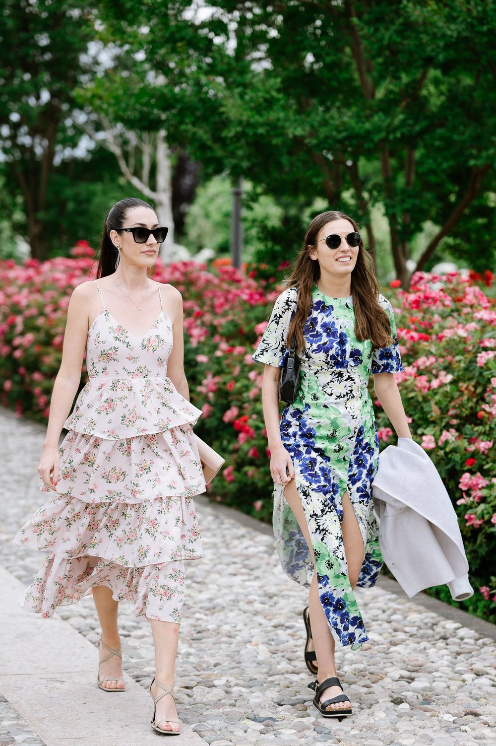 Two women in floral dresses walking along garden path wearing sunglasses.