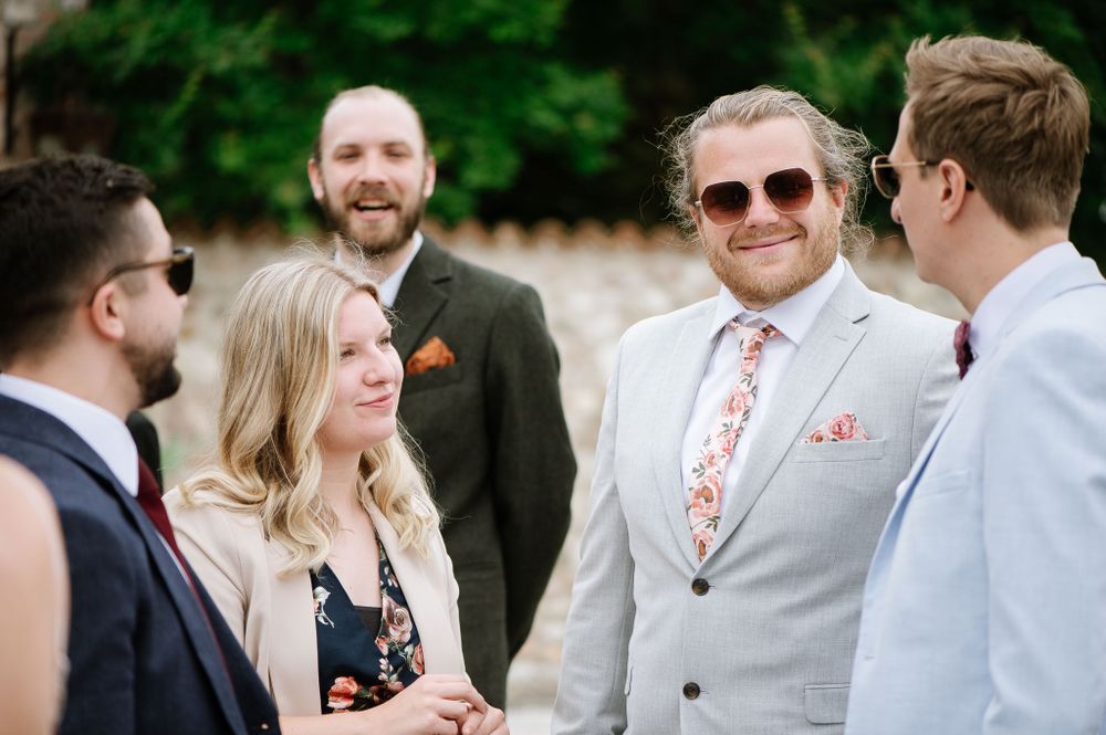 Wedding guests gather in casual conversation outdoors wearing formal attire and sunglasses.