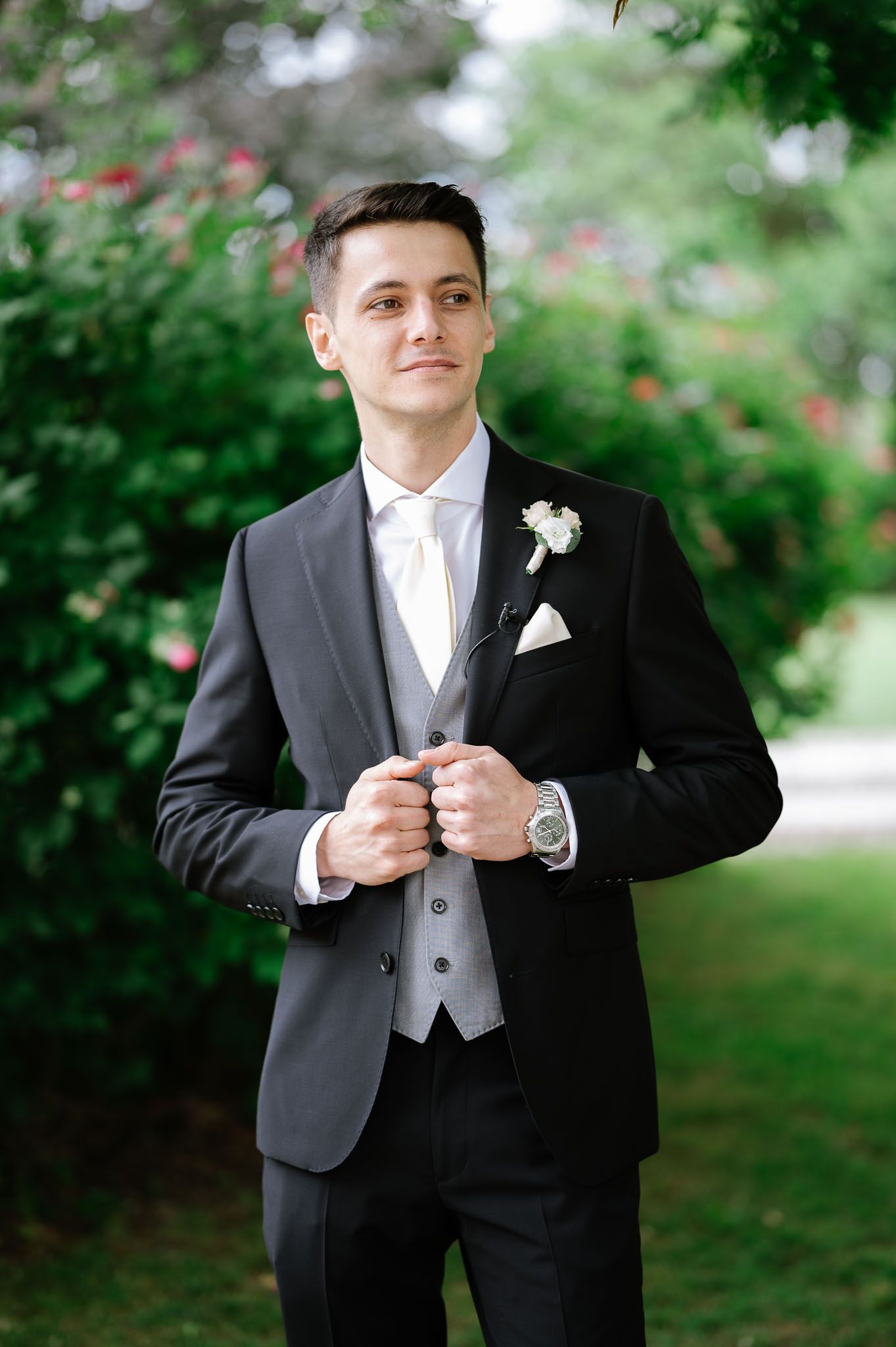 Groom adjusts his black tuxedo jacket while posing in front of lush green garden backdrop.