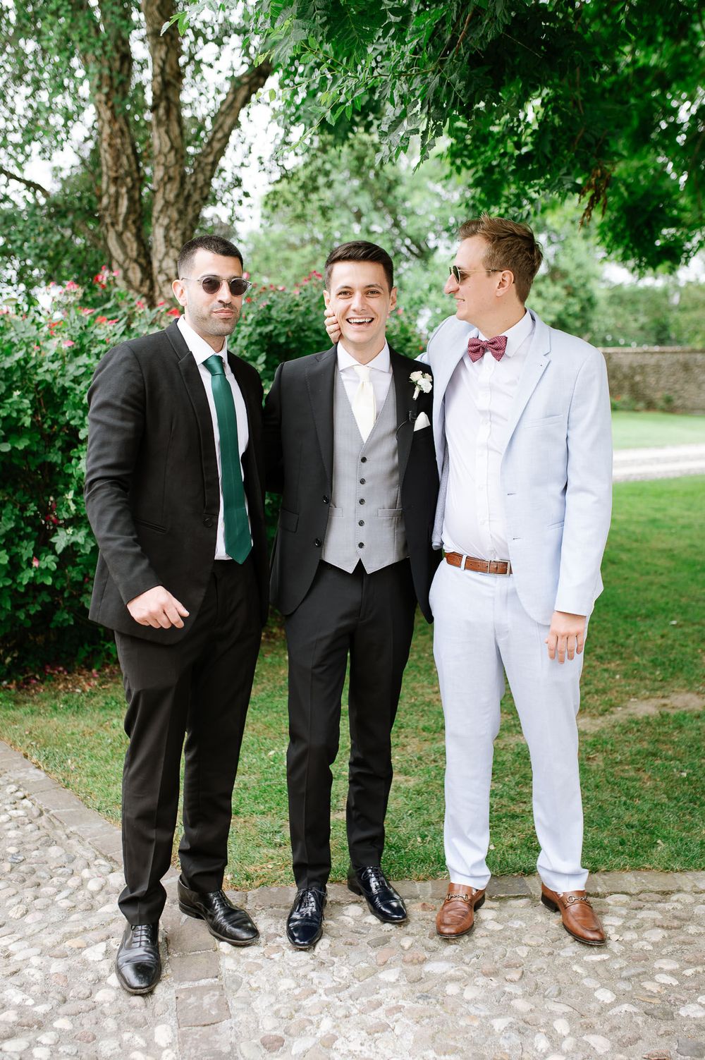 Three stylishly dressed men pose together at wedding, wearing contrasting suits and ties.