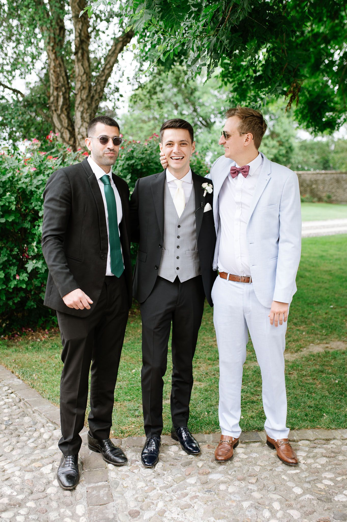 Three stylishly dressed men pose together at wedding, wearing contrasting suits and ties.