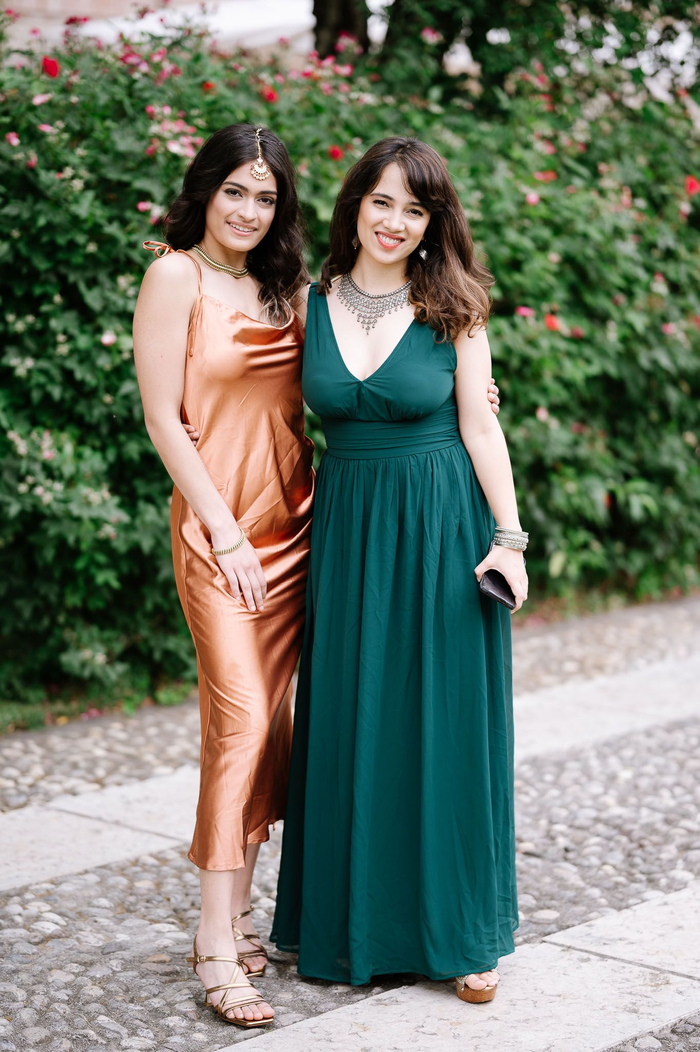 Two women in elegant dresses pose together against flowering garden backdrop.