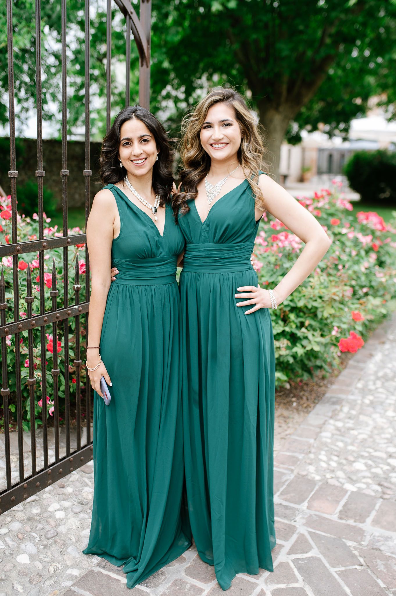 Two bridesmaids in matching emerald green formal gowns pose by rose garden.
