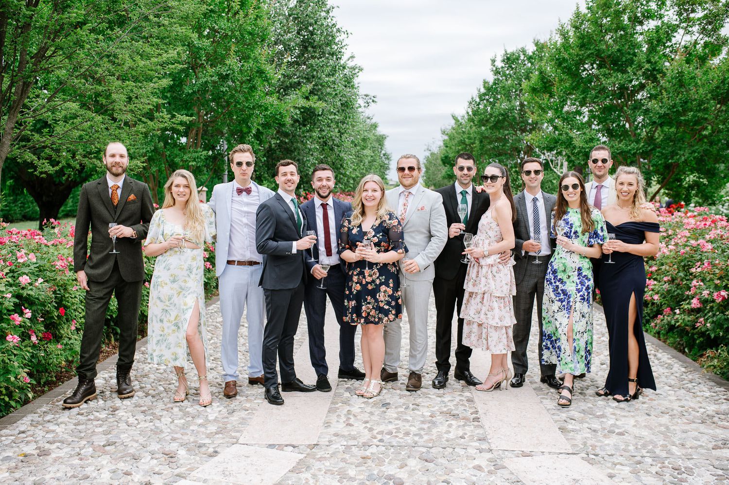 Wedding party group poses together on garden path surrounded by roses and greenery.