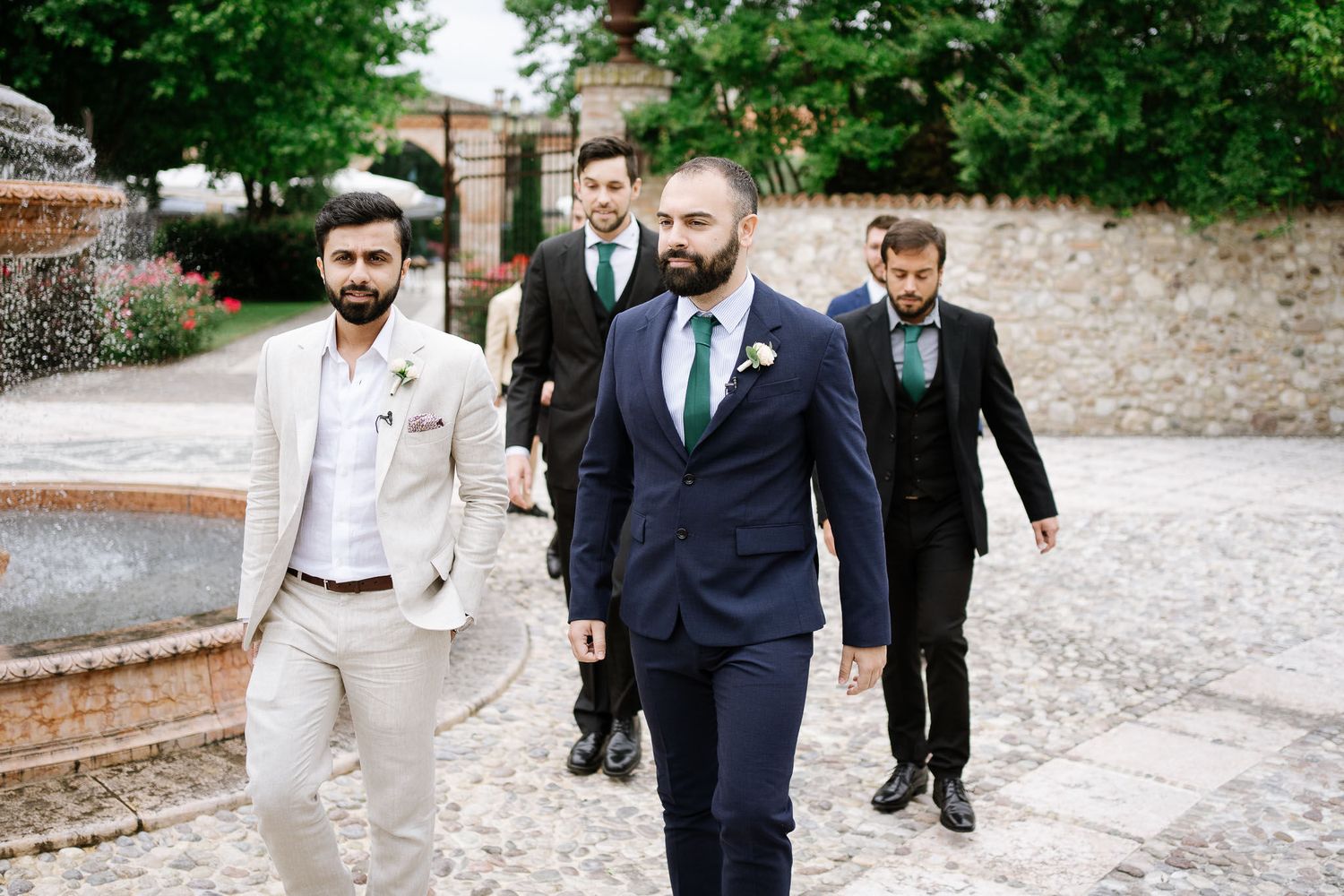 Wedding party members walk along a stone path at an outdoor venue wearing formal attire.