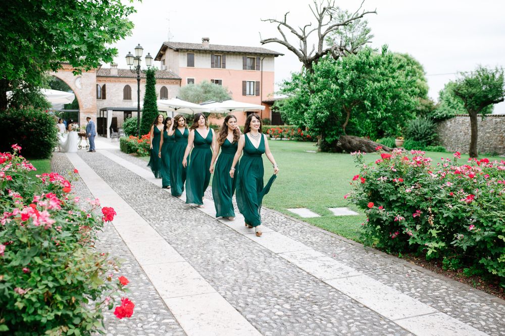Bridesmaids in matching green dresses walk along a garden path lined with pink roses.