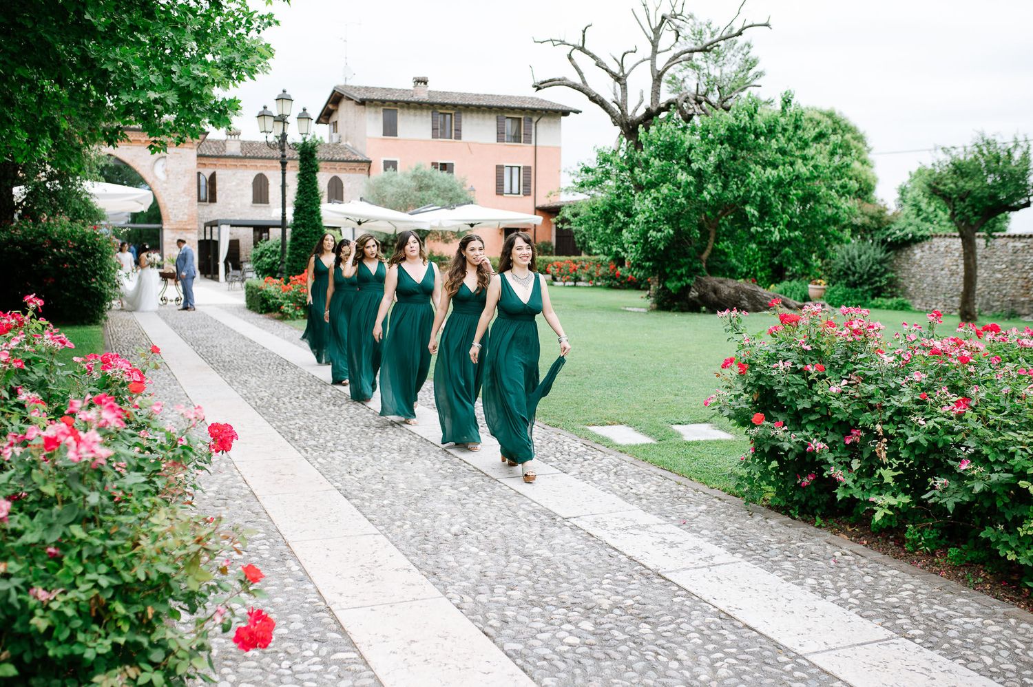 Bridesmaids in matching green dresses walk along a garden path lined with pink roses.