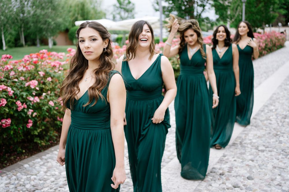Bridesmaids in emerald green dresses walking in line along a rose garden path.