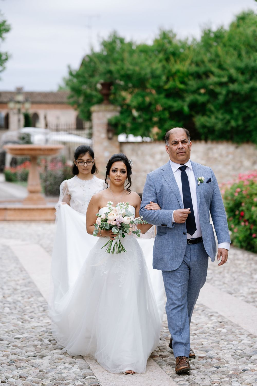 A bridal party walks along a stone pathway surrounded by roses and villa architecture.