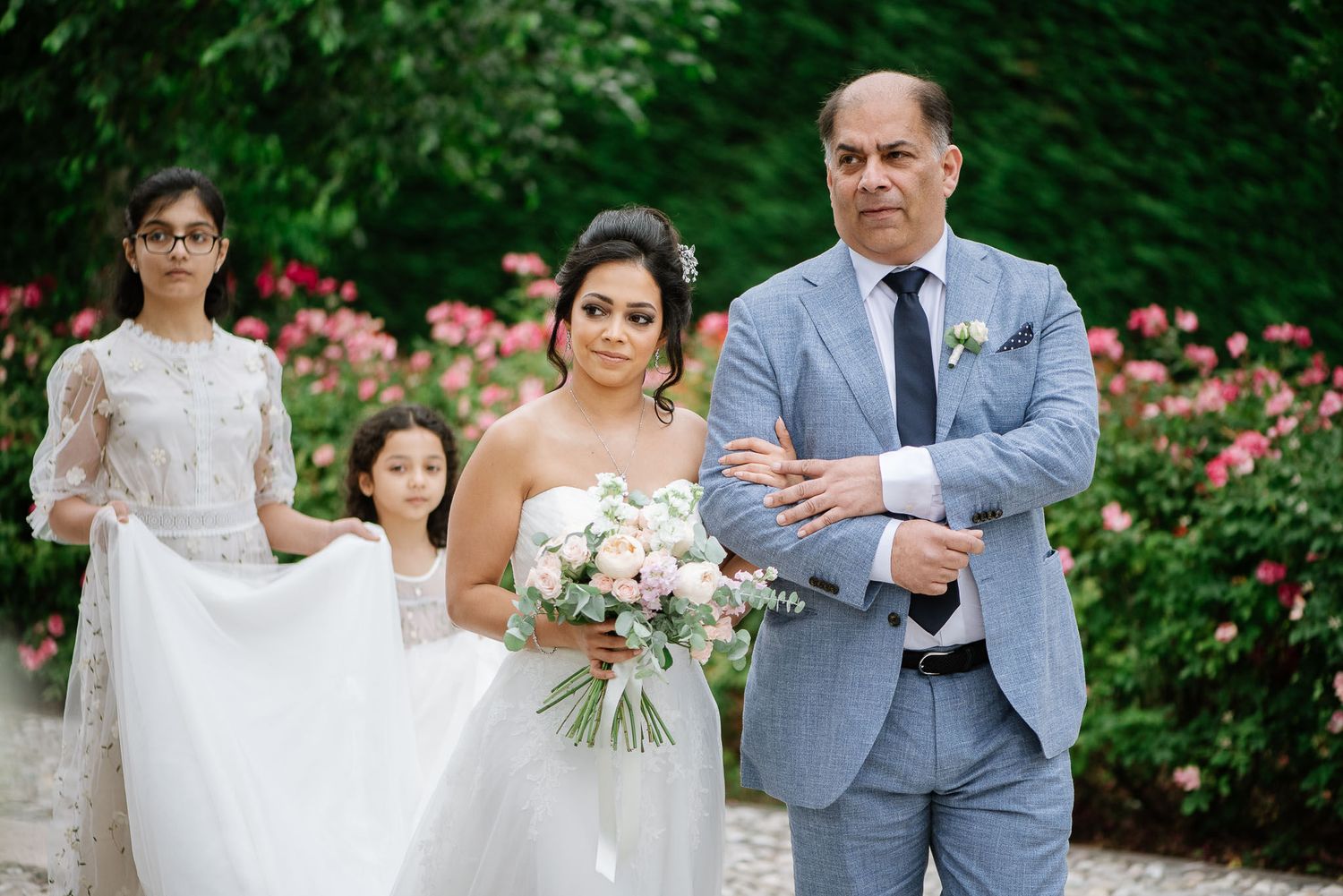 A wedding procession in a garden with pink flowers, featuring a bride in white gown and attendants.