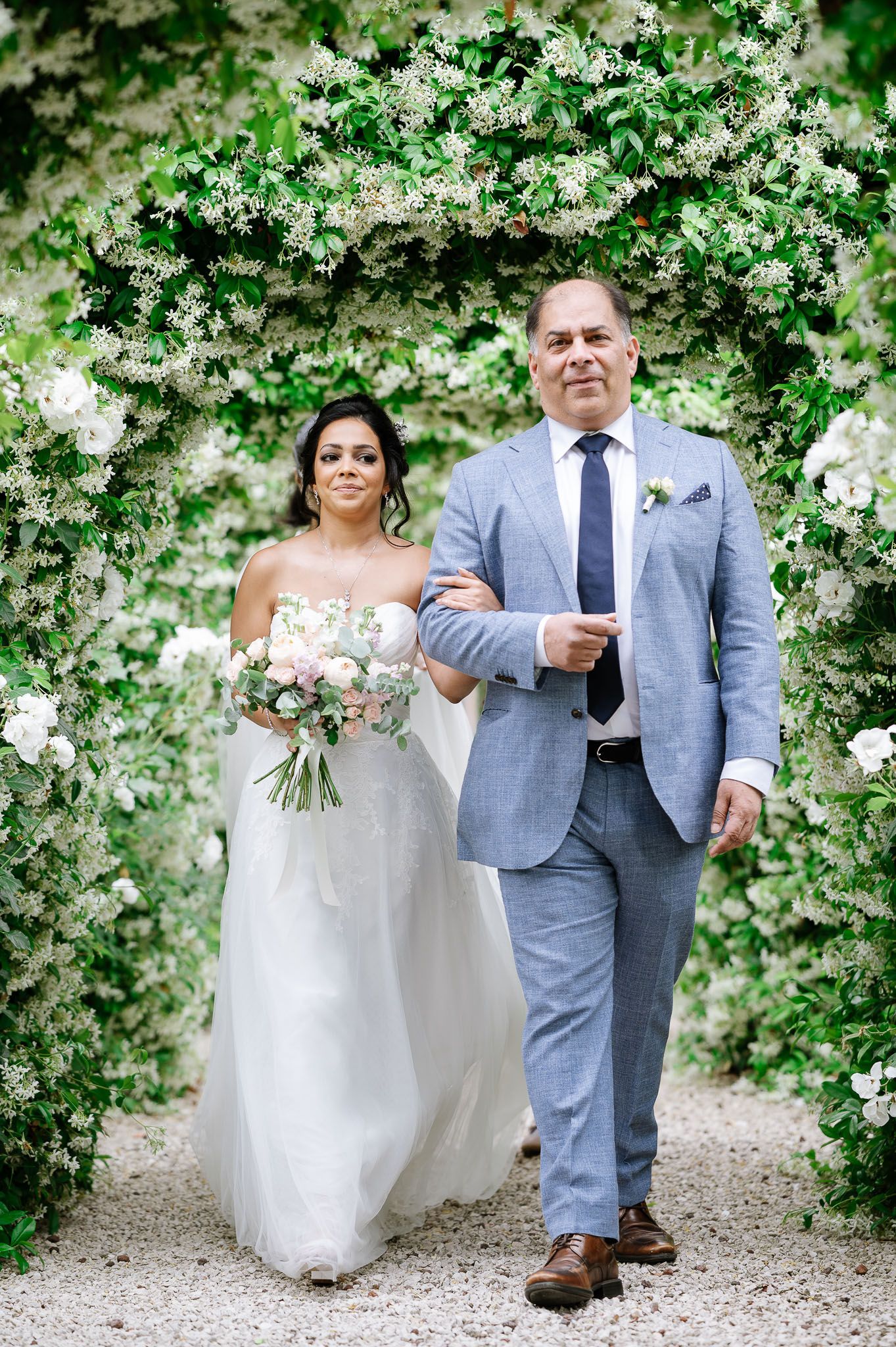 Wedding couple walks arm in arm under an archway of delicate white flowering vines.