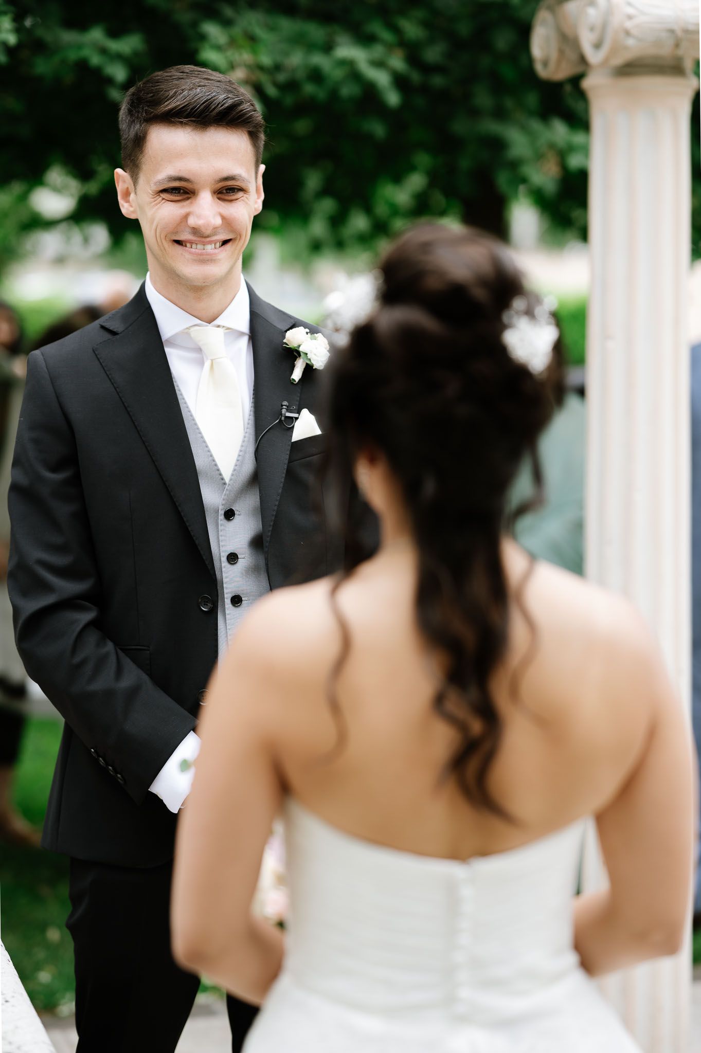A groom in formal attire smiles during an outdoor wedding ceremony.