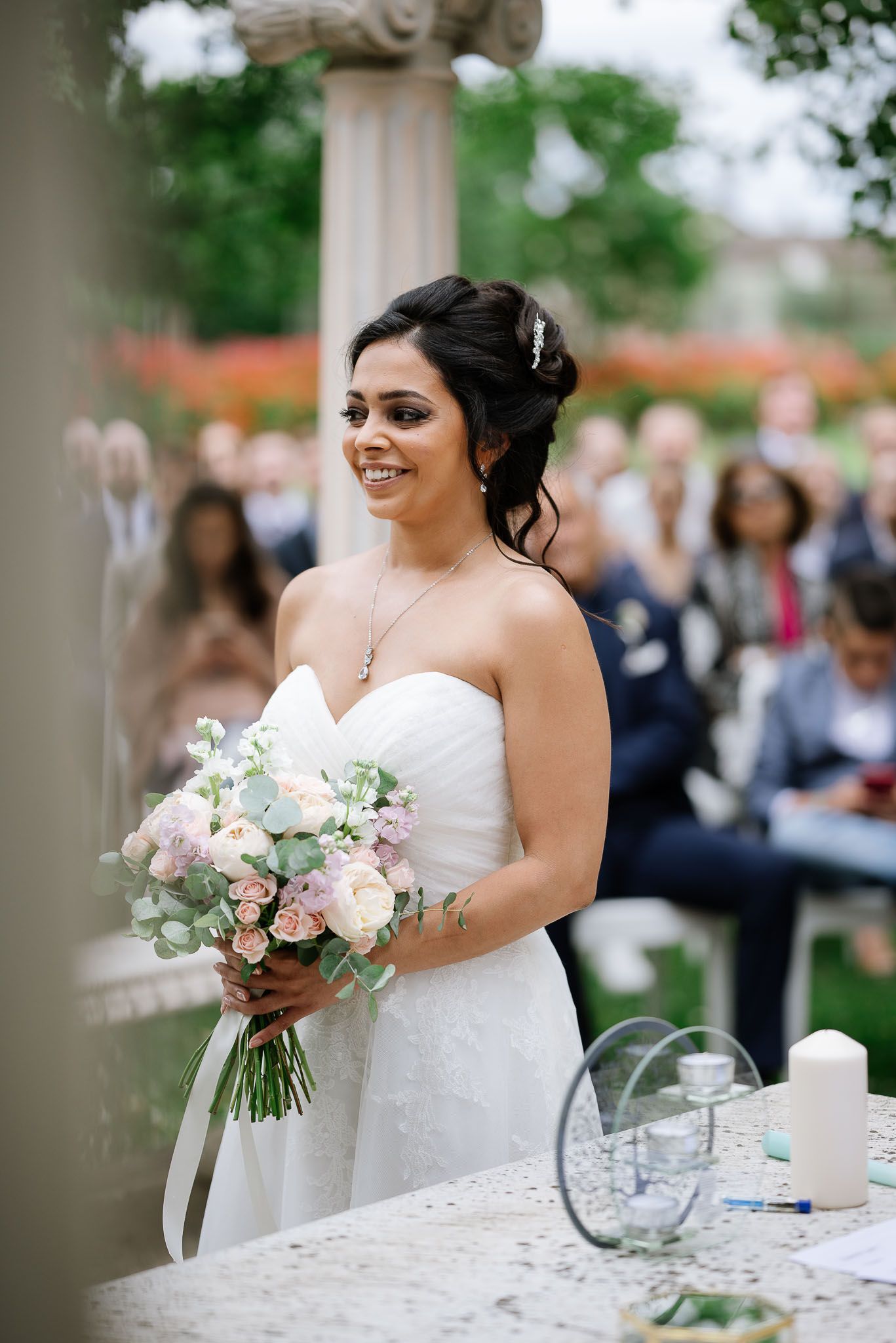 A bride in a strapless white gown holds a pink and white bouquet during an outdoor ceremony.