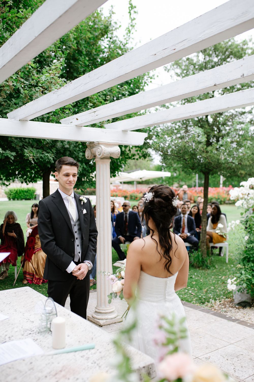 Outdoor wedding ceremony under a white pergola with classical columns and garden backdrop.