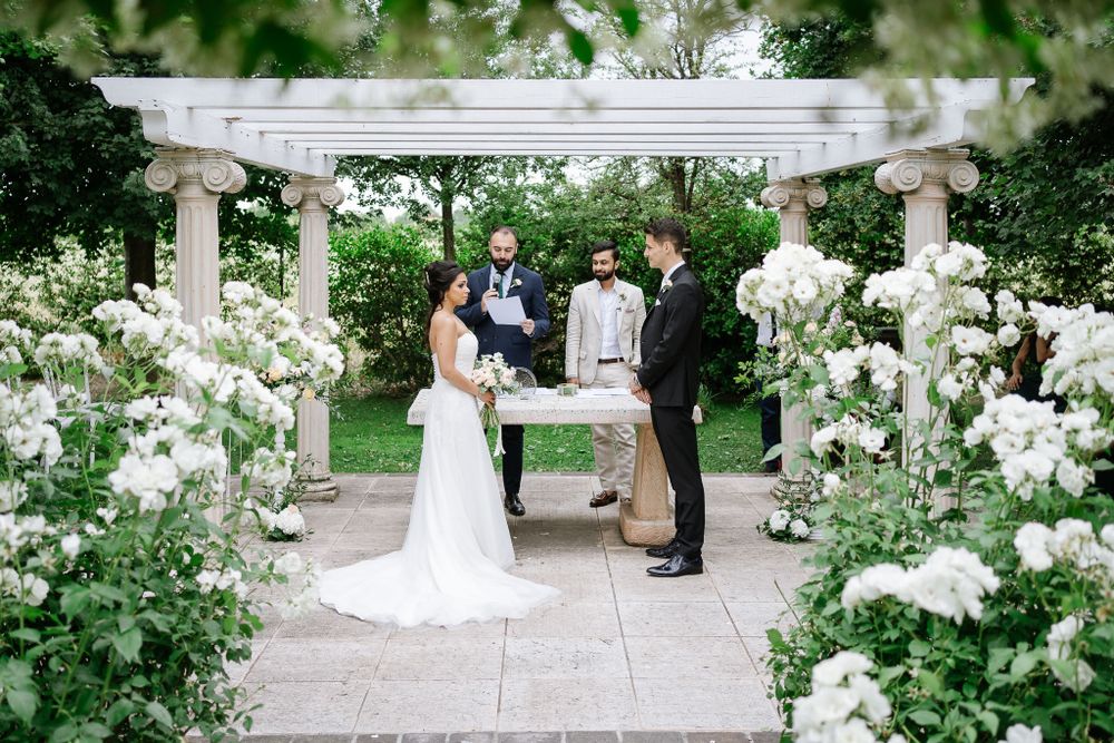 A wedding ceremony takes place under a white pergola surrounded by flowering gardens.
