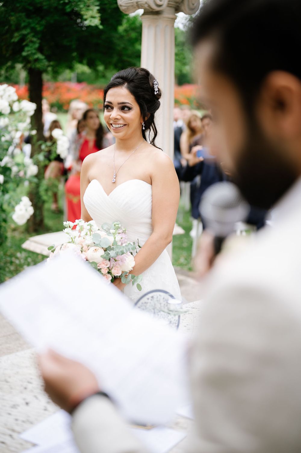 A bride smiles while holding her bouquet during an outdoor garden ceremony.