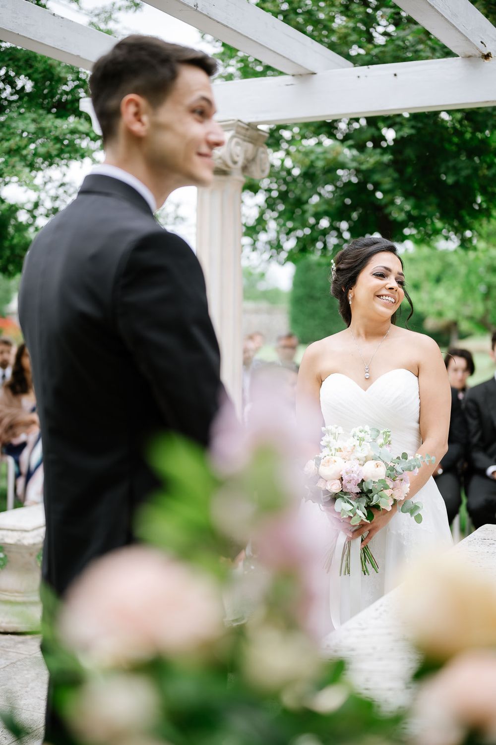 A bride in a strapless white gown shares a joyful moment with her partner under a garden pergola during their wedding ceremony.