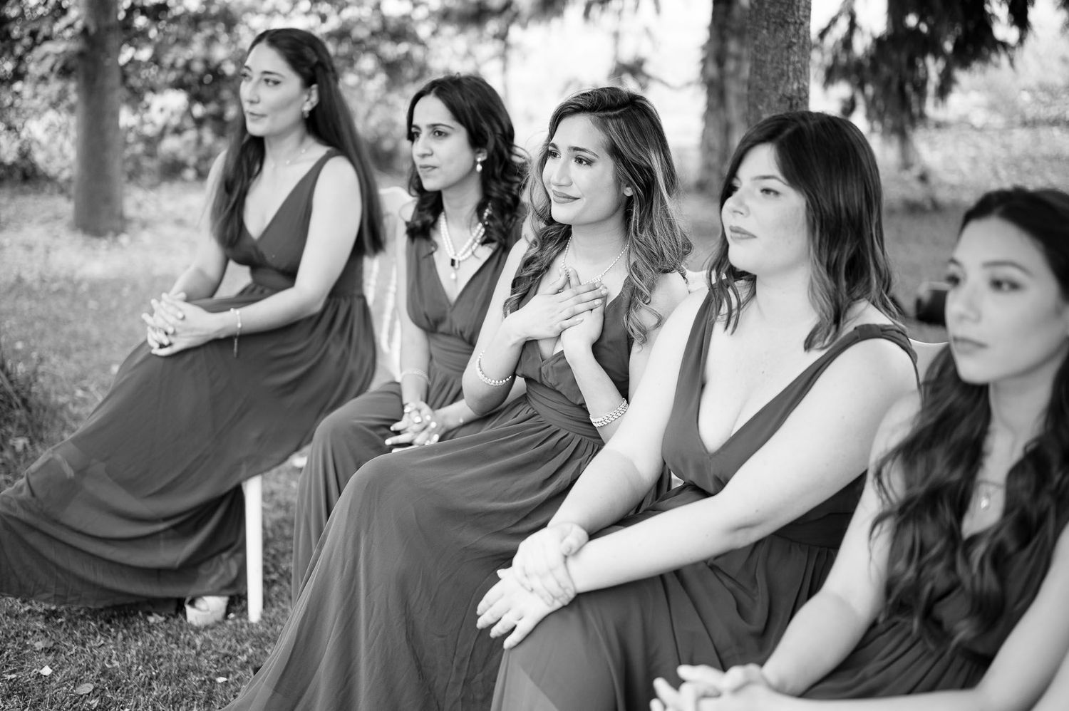 A row of bridesmaids in matching burgundy dresses sit together during an outdoor wedding ceremony.