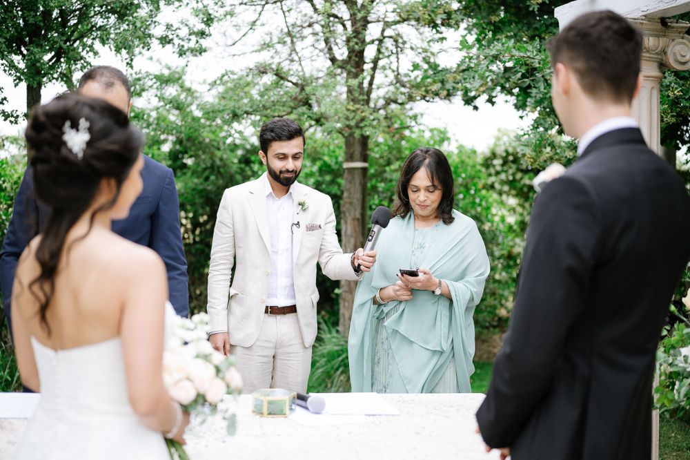 An outdoor wedding ceremony takes place under green trees with an officiant leading the service.
