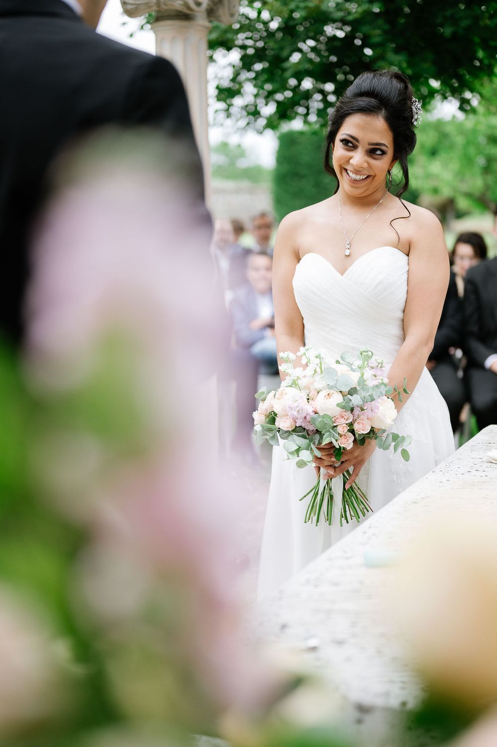 A bride in a sweetheart neckline dress holds a delicate pink and white bouquet during an outdoor ceremony.