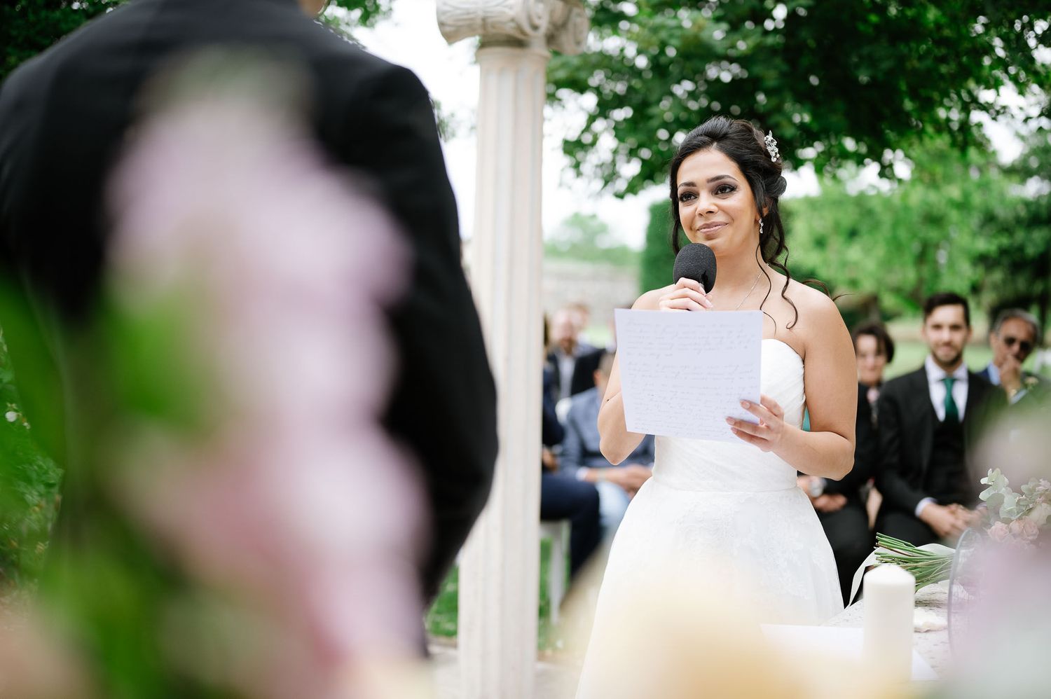 A bride reads her vows from a paper during an outdoor garden wedding ceremony.