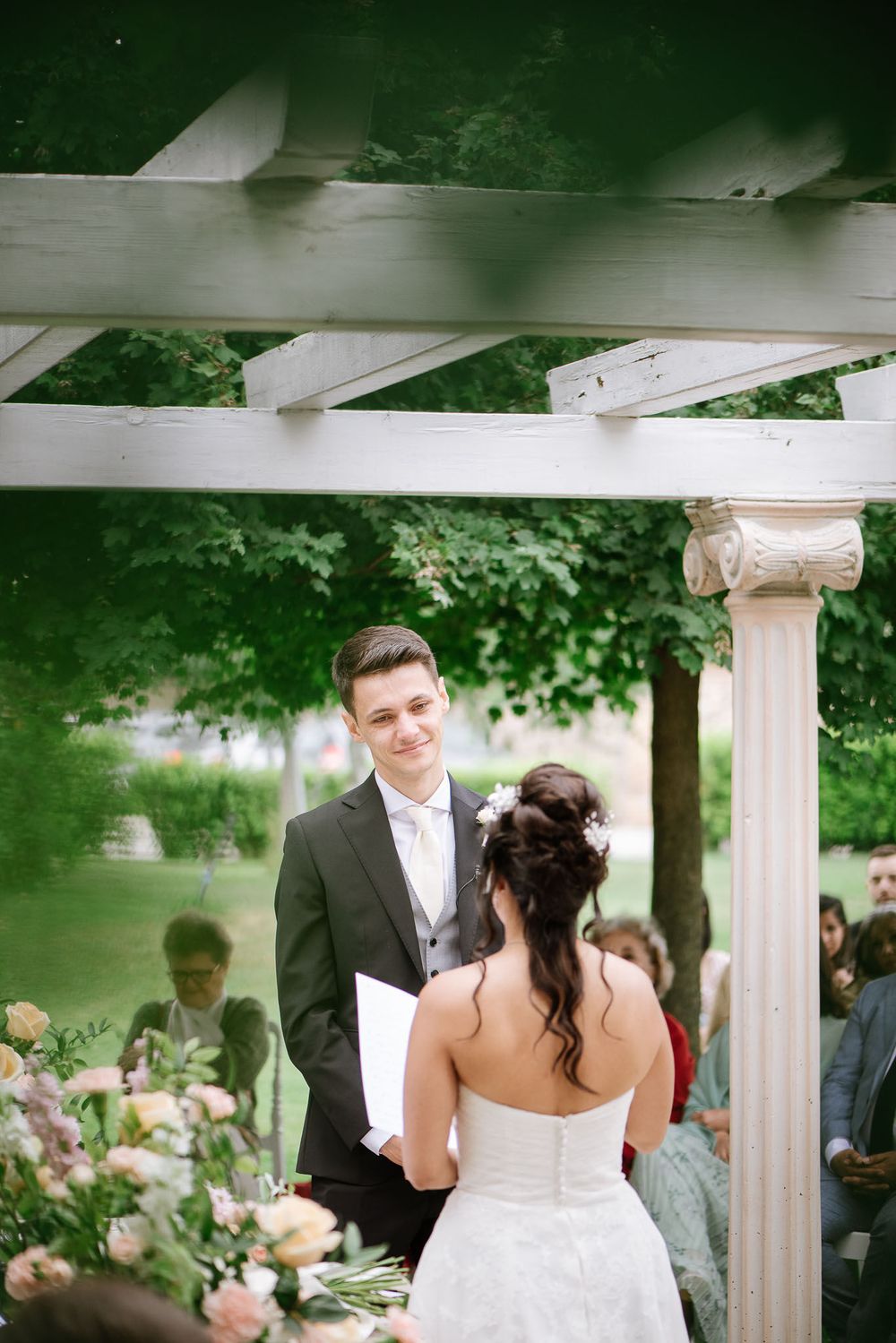 A couple exchanges vows beneath a white pergola surrounded by lush garden greenery.