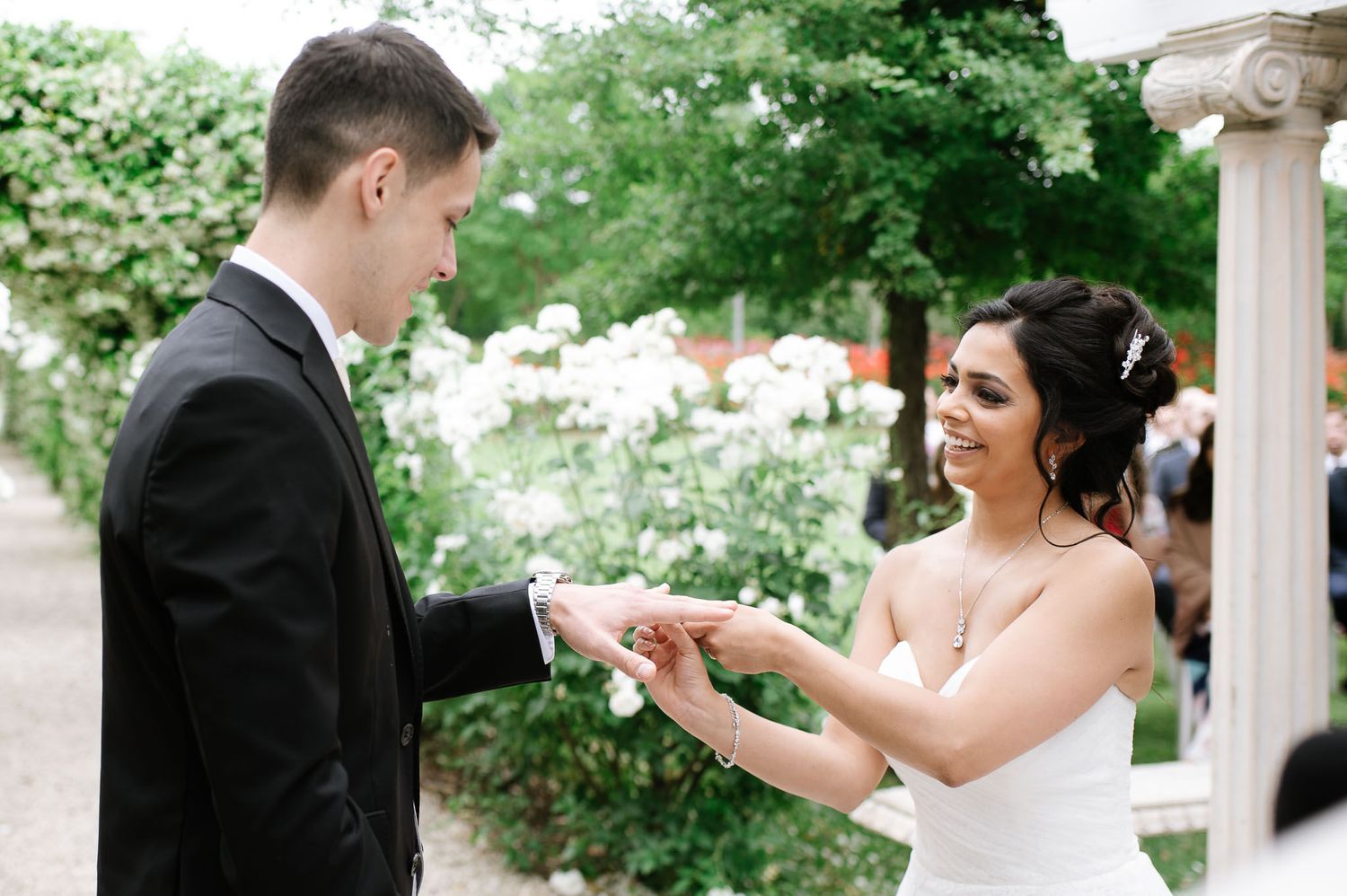 A couple exchanges wedding rings in a garden setting surrounded by blooming flowers.