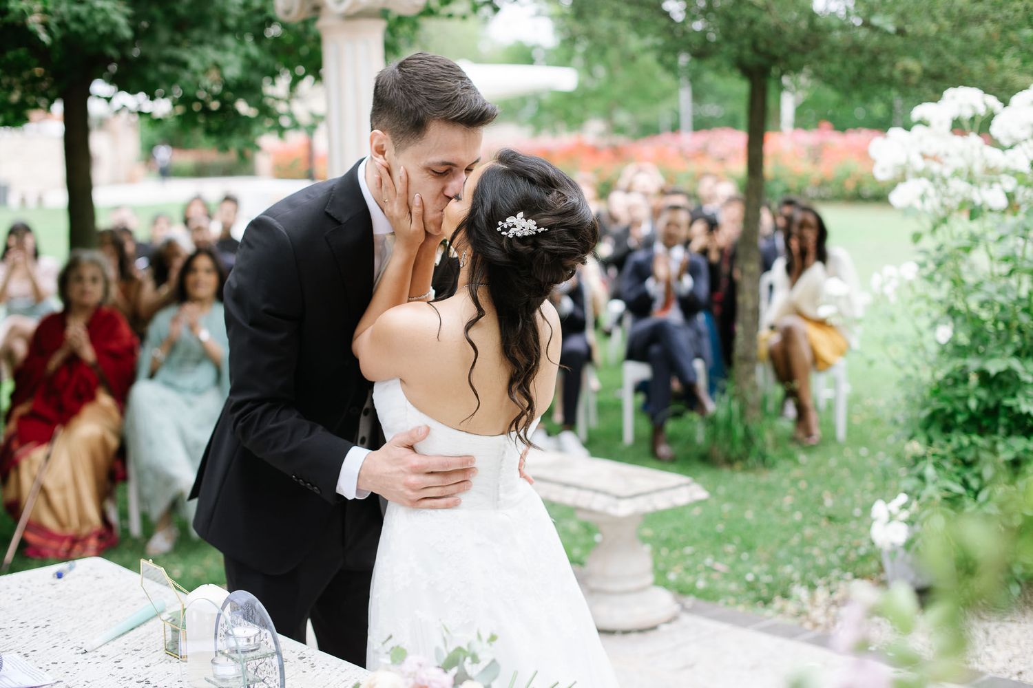 A newlywed couple shares a romantic kiss during their garden wedding ceremony surrounded by guests.