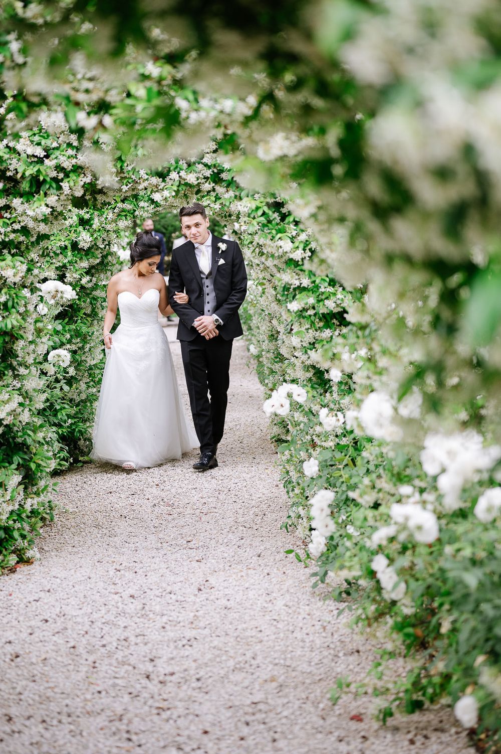 A couple walks down a garden path lined with blooming white flowers on their wedding day.