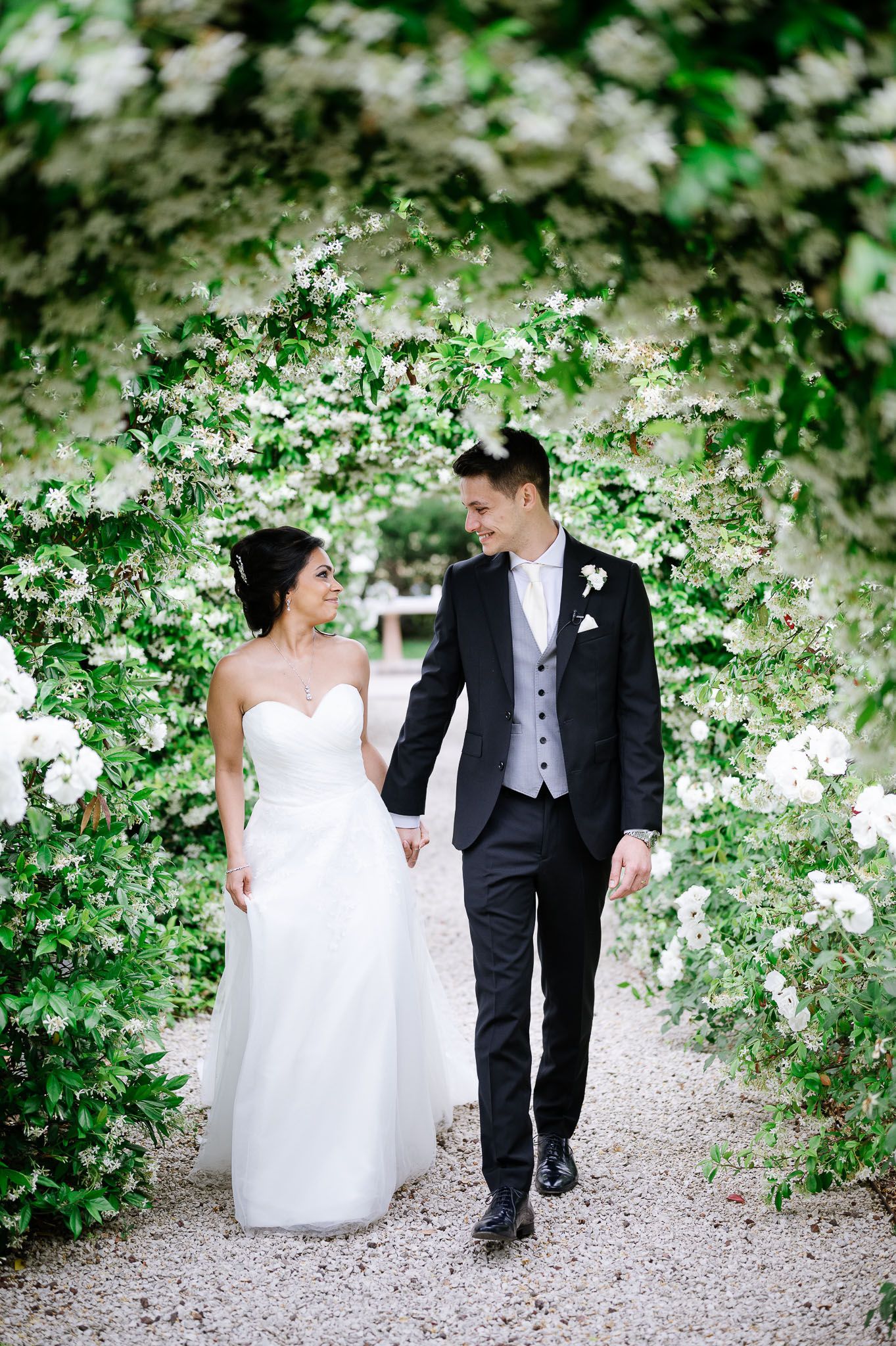 A bride and groom stroll through a garden pathway surrounded by blooming white flowers.