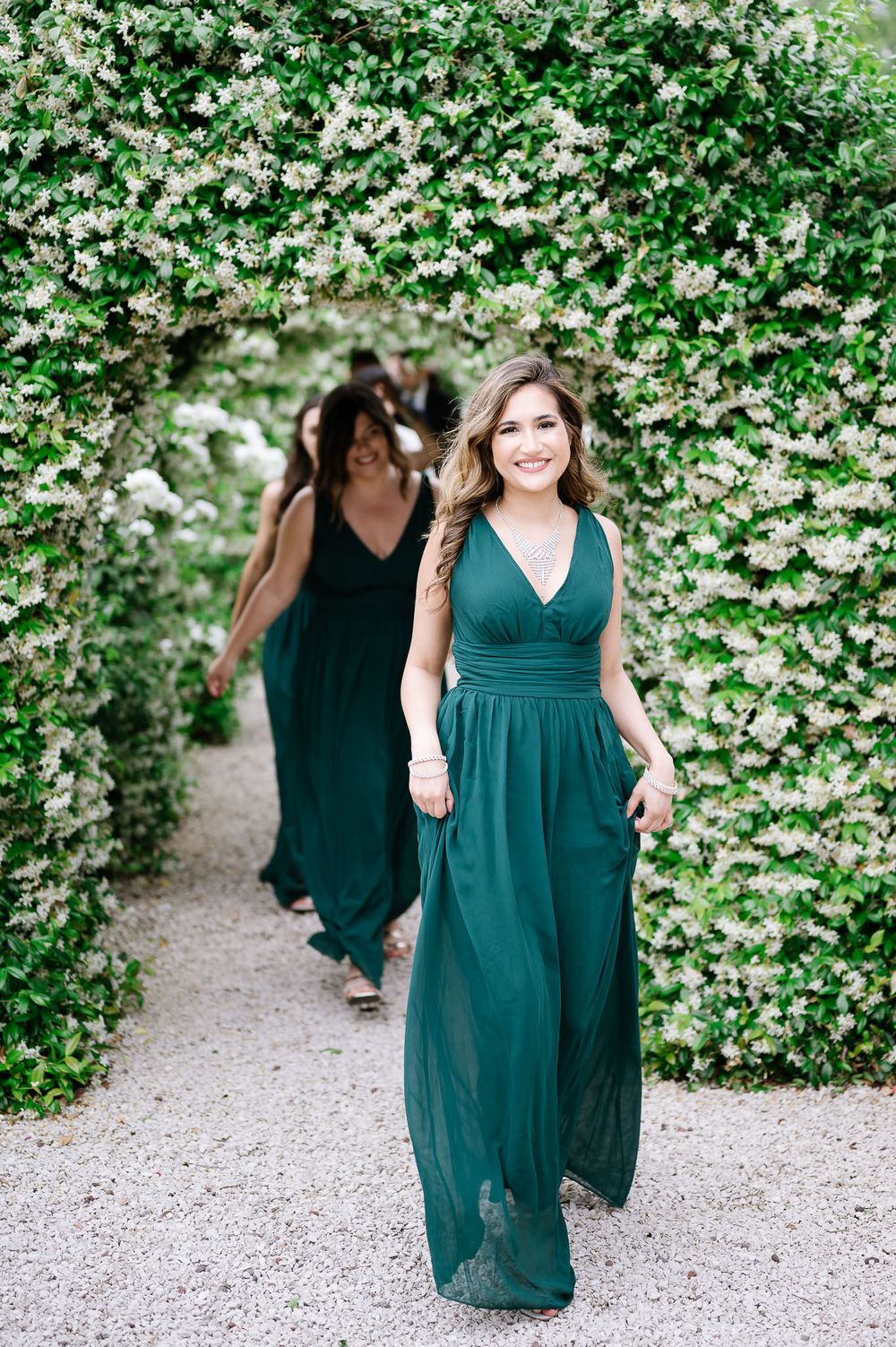 Bridesmaids in emerald green dresses walk through a floral archway.