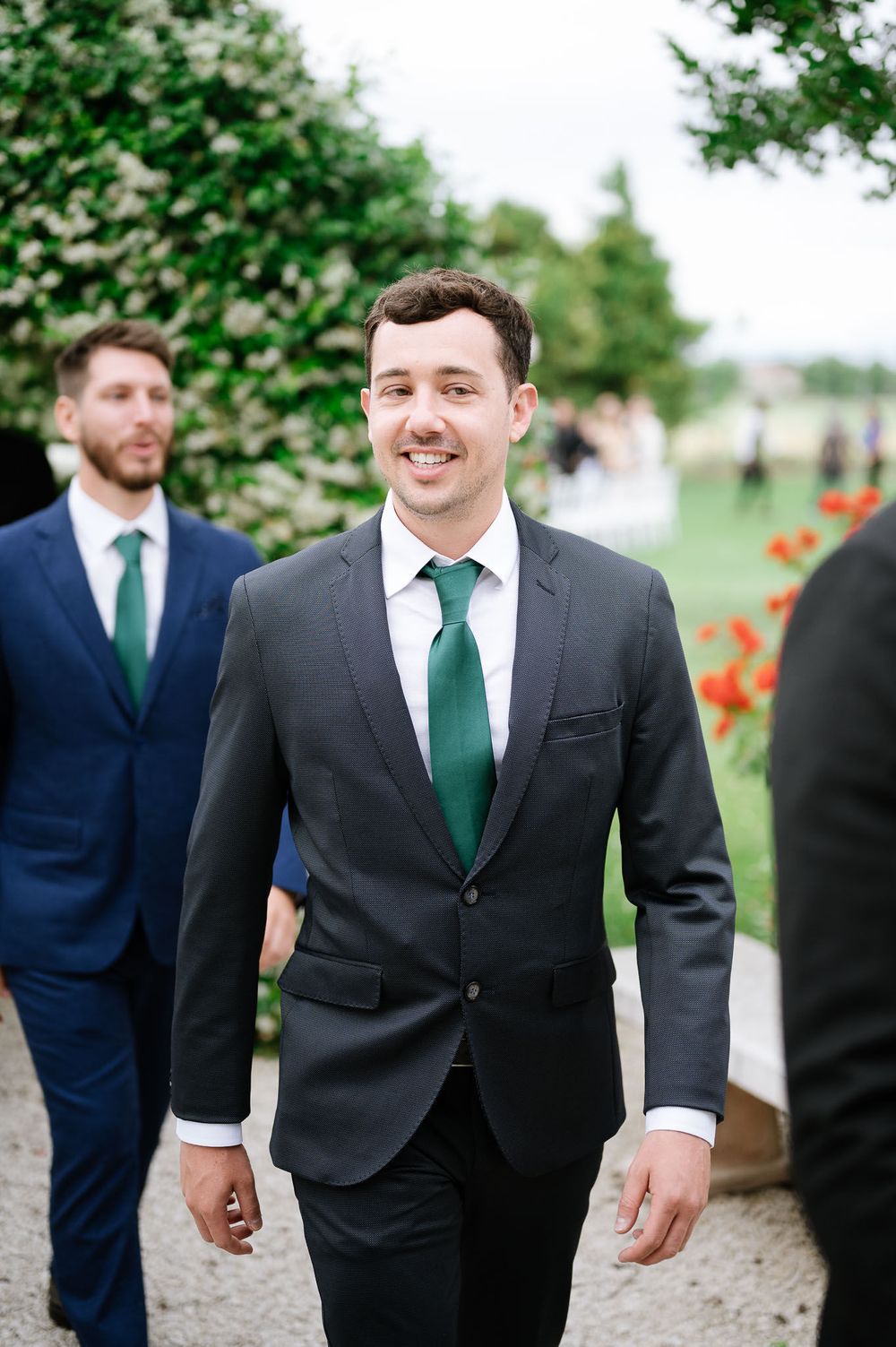 Wedding guest in dark suit and green tie walks along garden path.