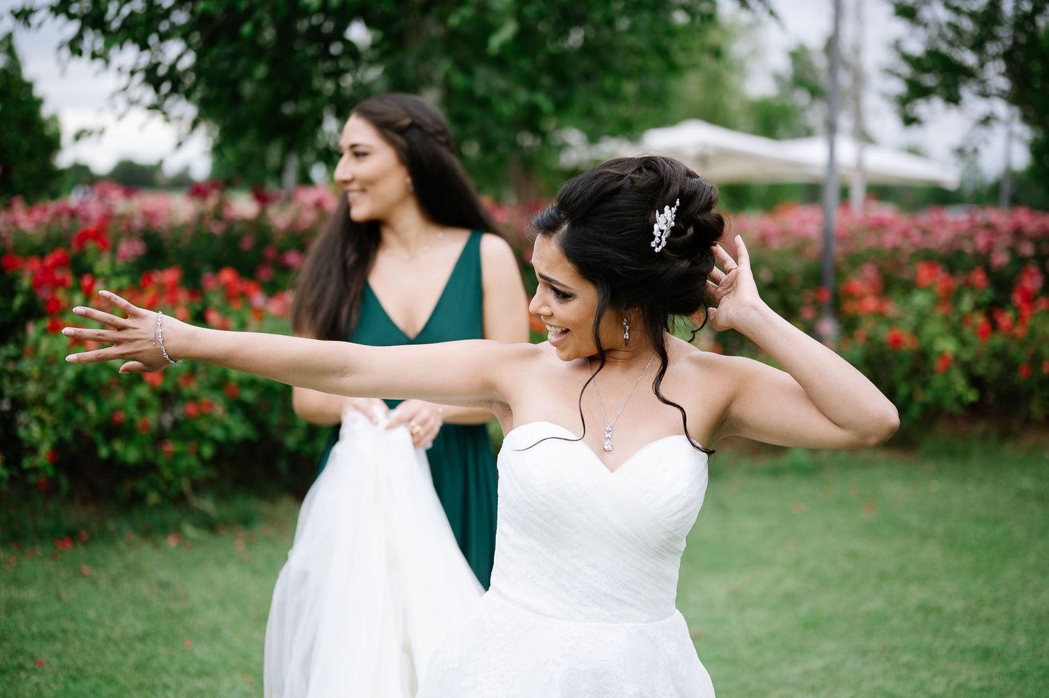 Two women in wedding attire celebrate joyfully in a garden with red flowers in the background.