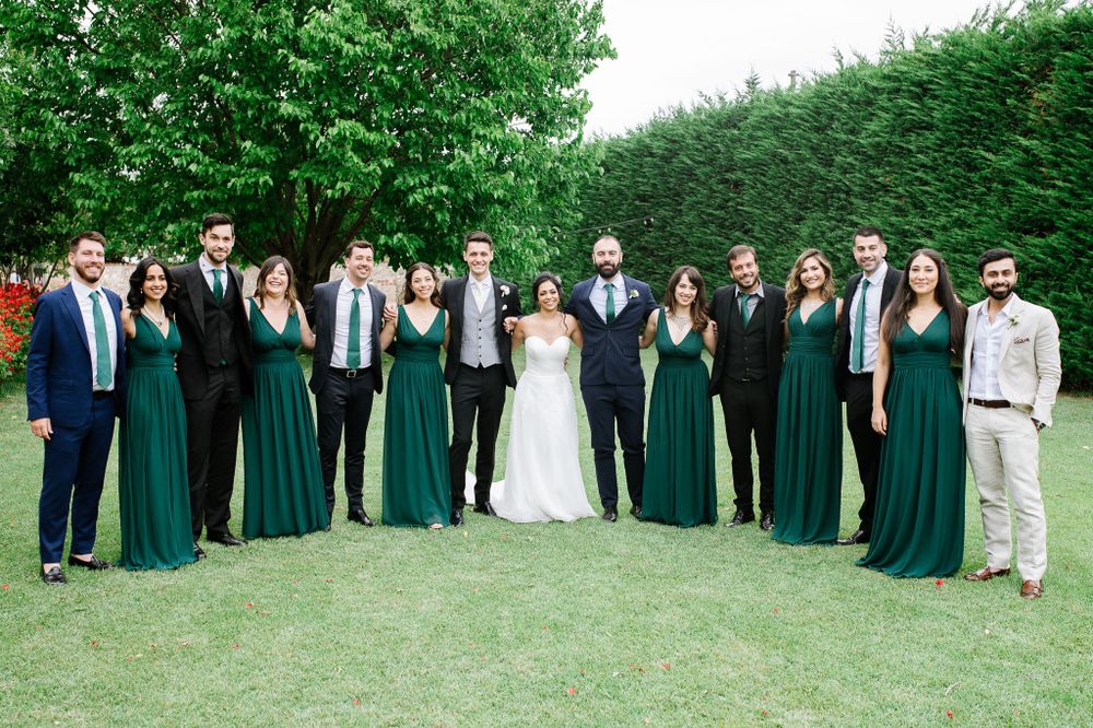 Full wedding party in emerald green and formal attire poses on grass for group photo.