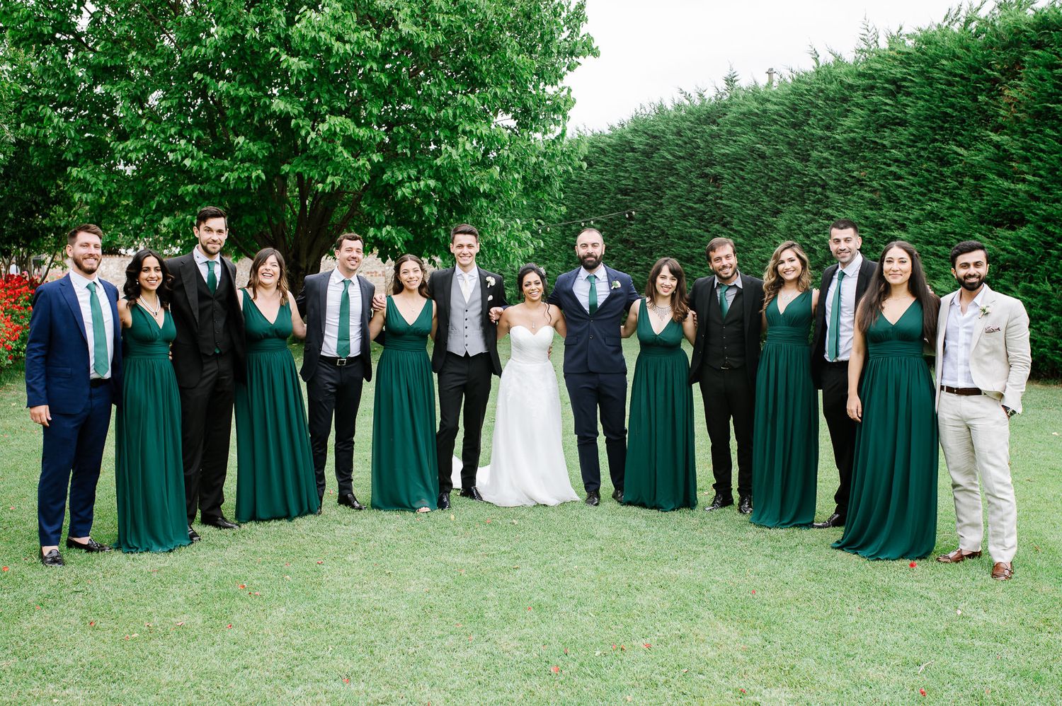 Full wedding party in emerald green and formal attire poses on grass for group photo.