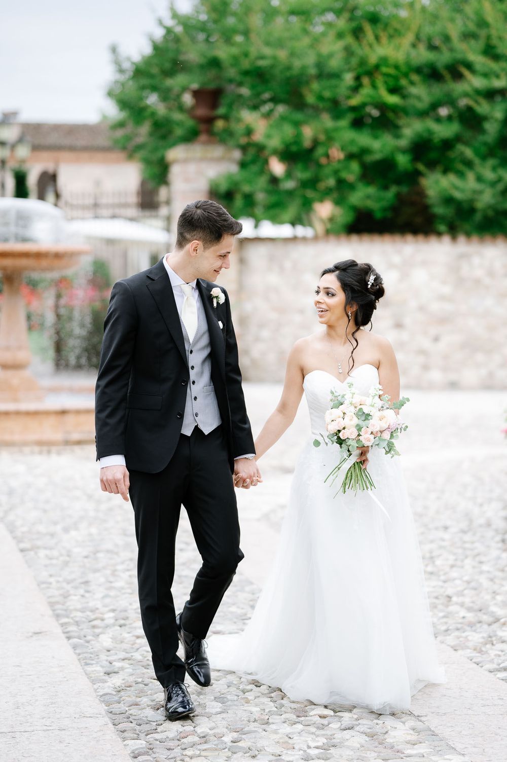 Newlyweds walk hand in hand on cobblestone path with bride holding delicate bouquet.