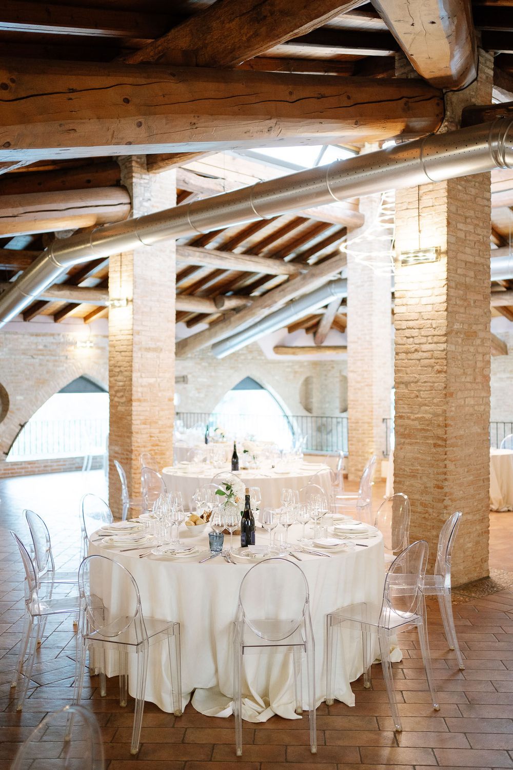 Elegant wedding reception table setting under rustic wooden beams with clear ghost chairs.
