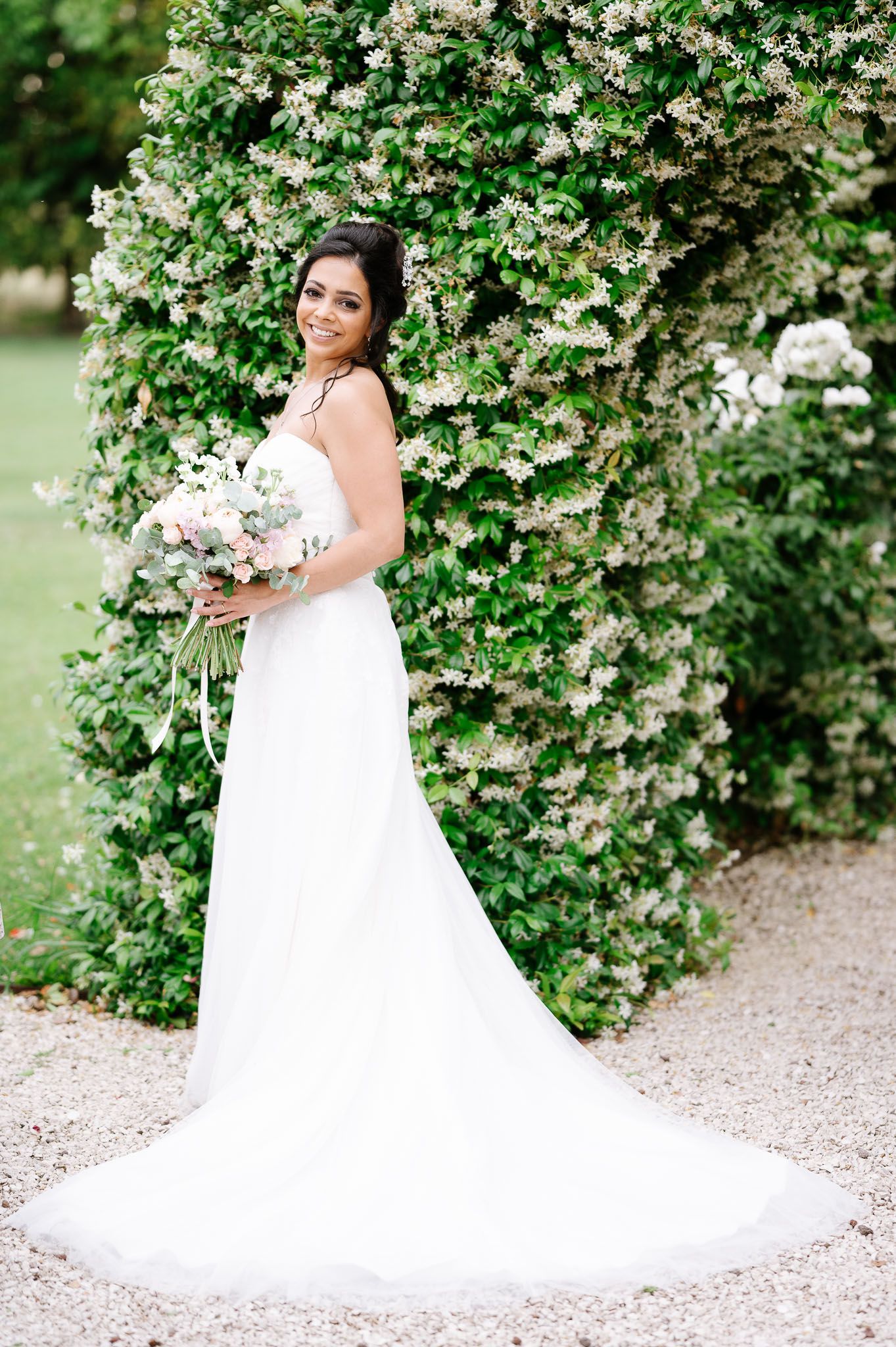 Bride in flowing white wedding dress poses elegantly against blooming white floral backdrop in garden setting.