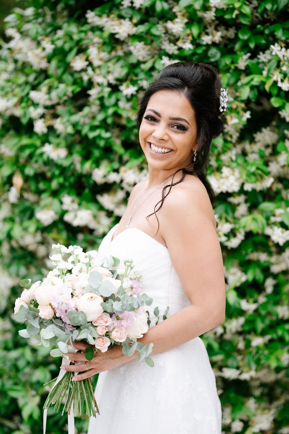 Joyful bride holds pastel bouquet while posing against flowering garden backdrop.