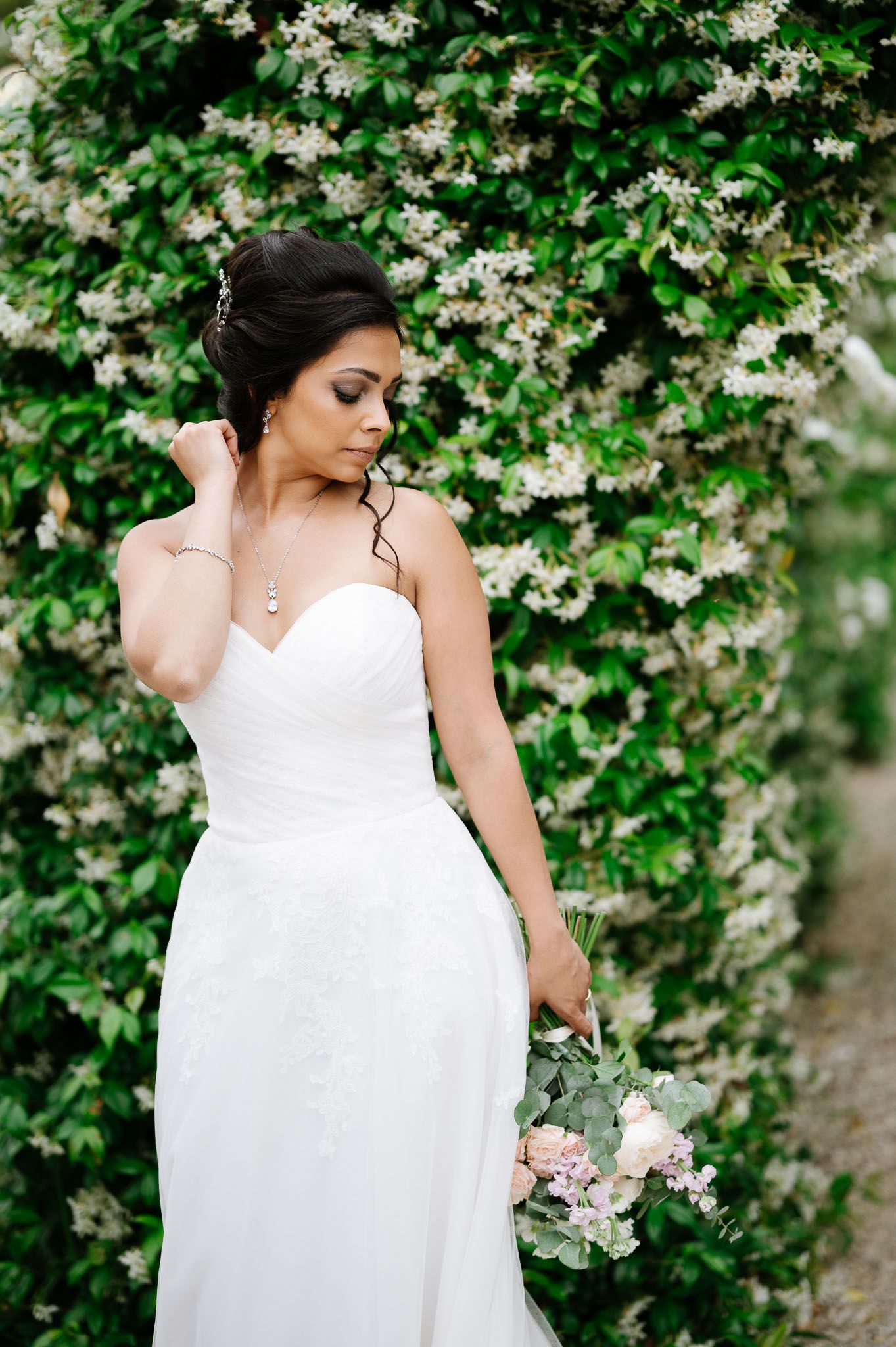 Elegant bride in white gown poses against flowering garden wall with delicate bouquet.