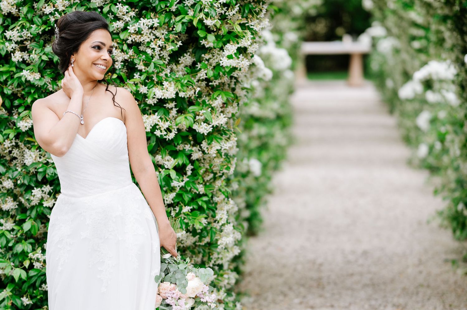 Bride in classic white dress poses along garden path lined with flowering white blooms.