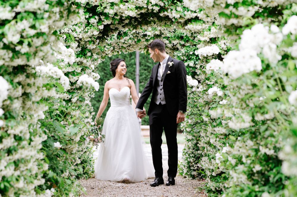 Newlyweds walking hand in hand through romantic garden arch covered in white flowers.