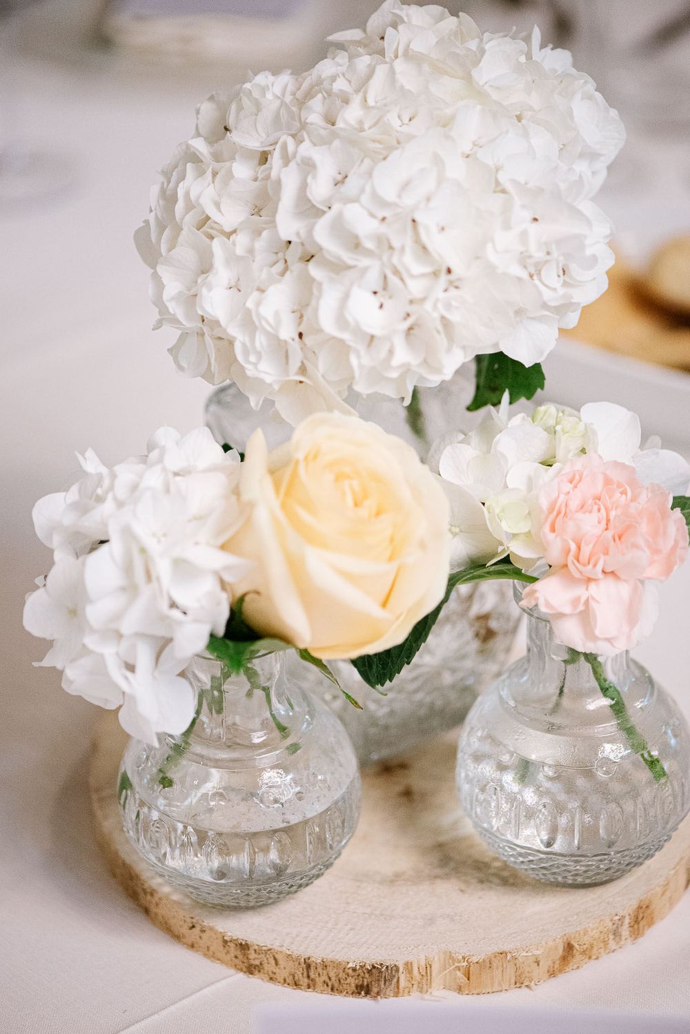 Elegant floral centerpiece with white hydrangeas, cream rose and pink carnation in crystal vases on wooden slice.