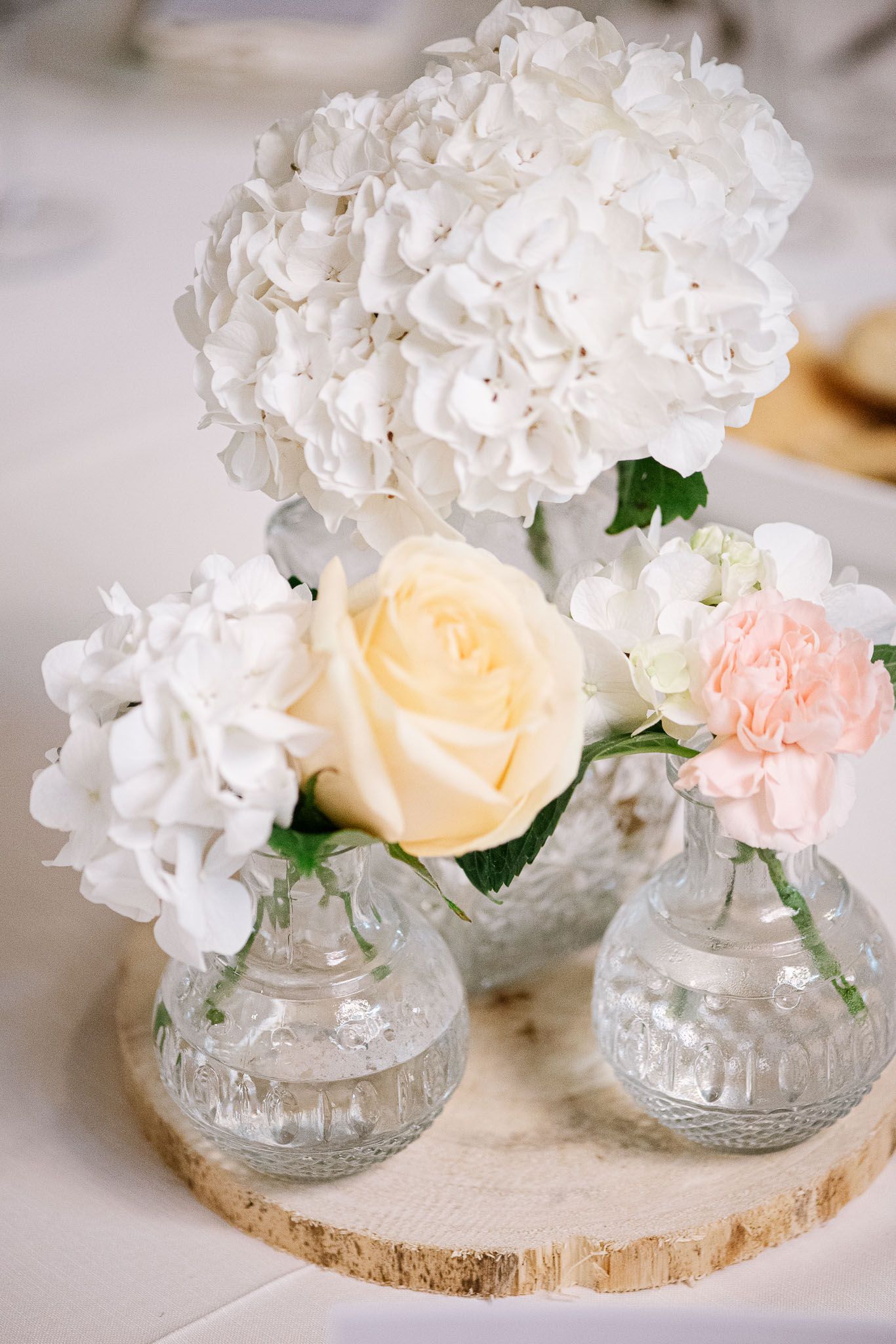 Elegant floral centerpiece with white hydrangeas, cream rose and pink carnation in crystal vases on wooden slice.