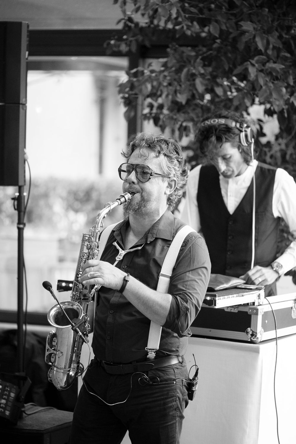 Black and white photo of saxophone musician performing with DJ in background.