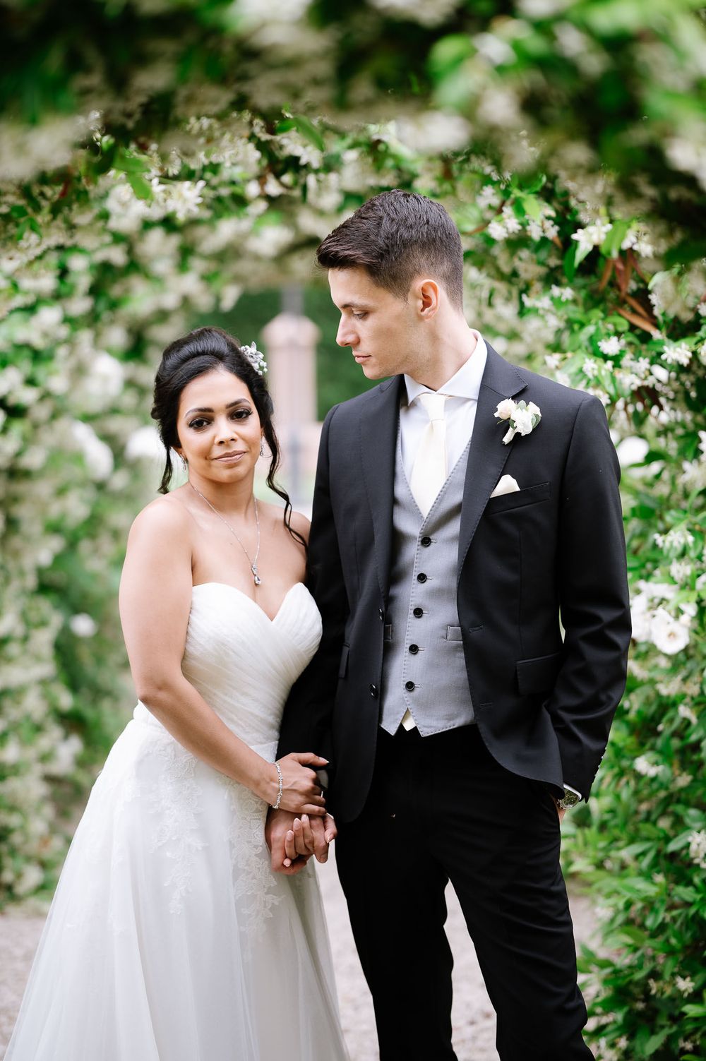 Elegant wedding couple poses among lush greenery and white flowers in garden setting.