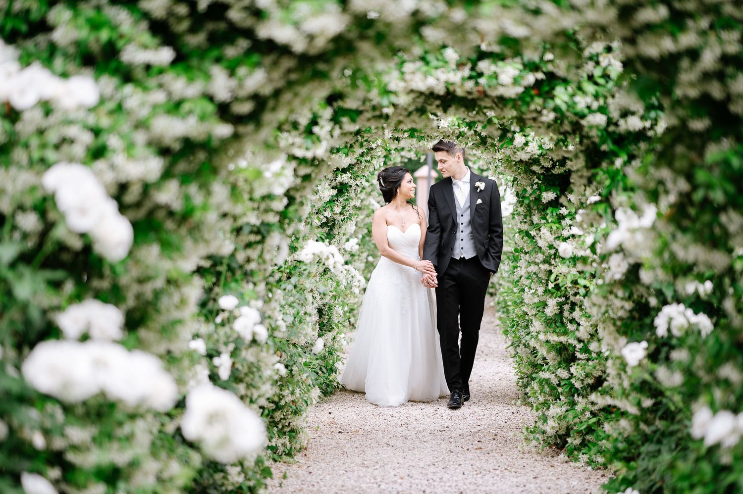 Romantic wedding photo of couple walking through flower-covered garden arch.