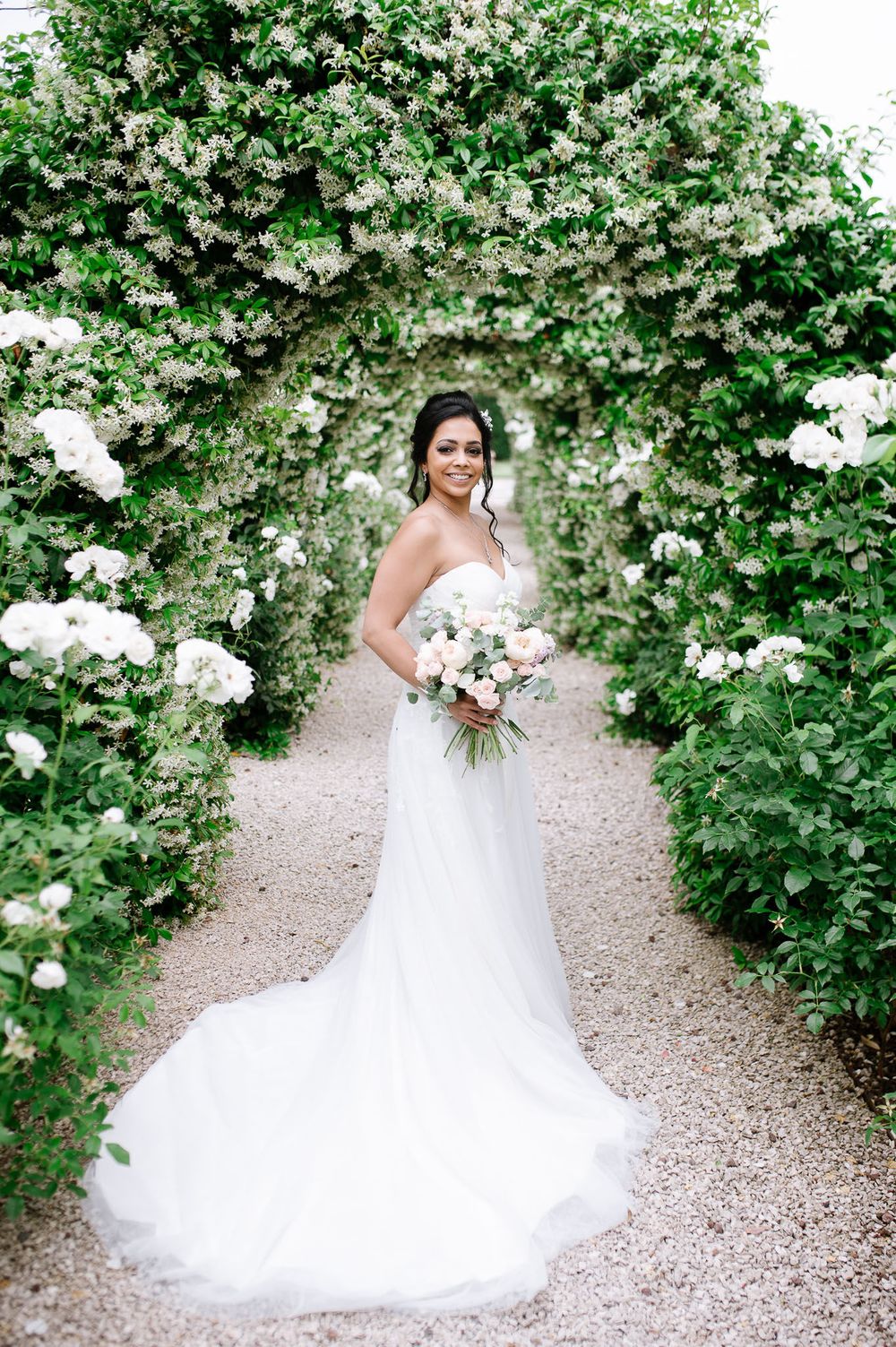 Bride in flowing white gown poses under floral garden arch holding bouquet.