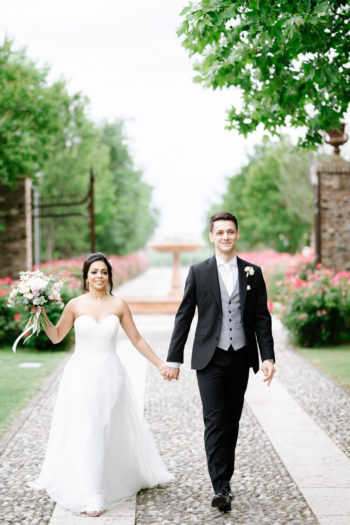 A couple walking hand in hand along a garden pathway lined with blooming flowers.