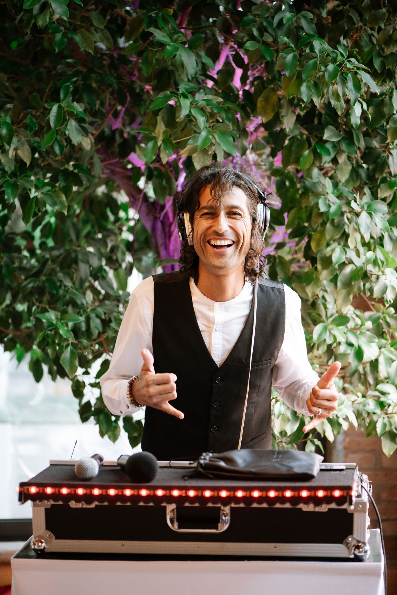 A DJ performs enthusiastically behind turntables under green foliage with purple accents.